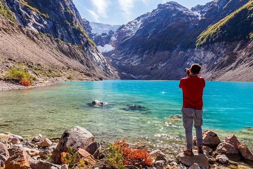 Un hombre con camisa roja fotografía un lago turquesa rodeado de altas montañas nevadas.