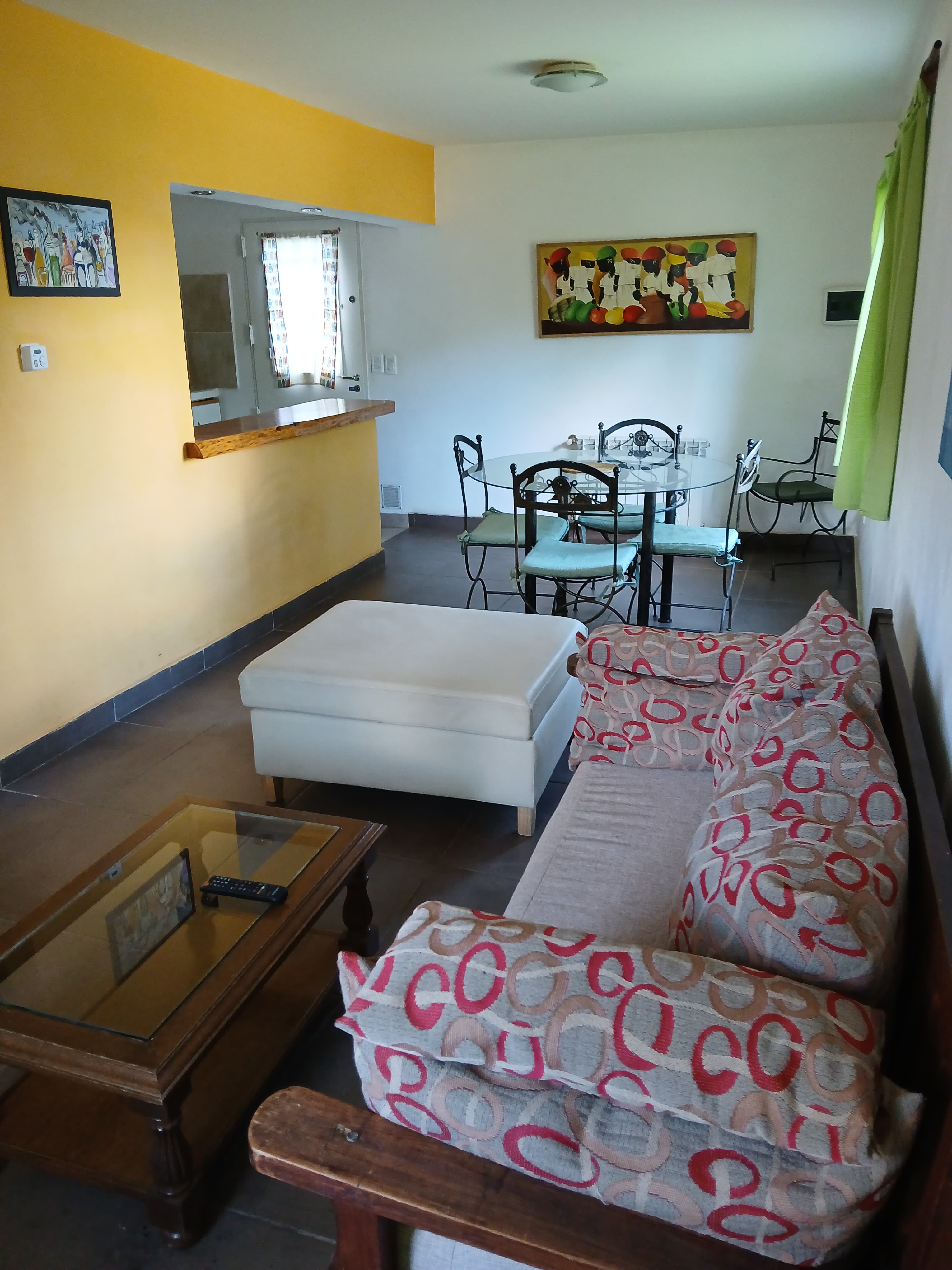 Sala con sofá de círculos rojos, otomana blanca, mesa de vidrio y pared amarilla.