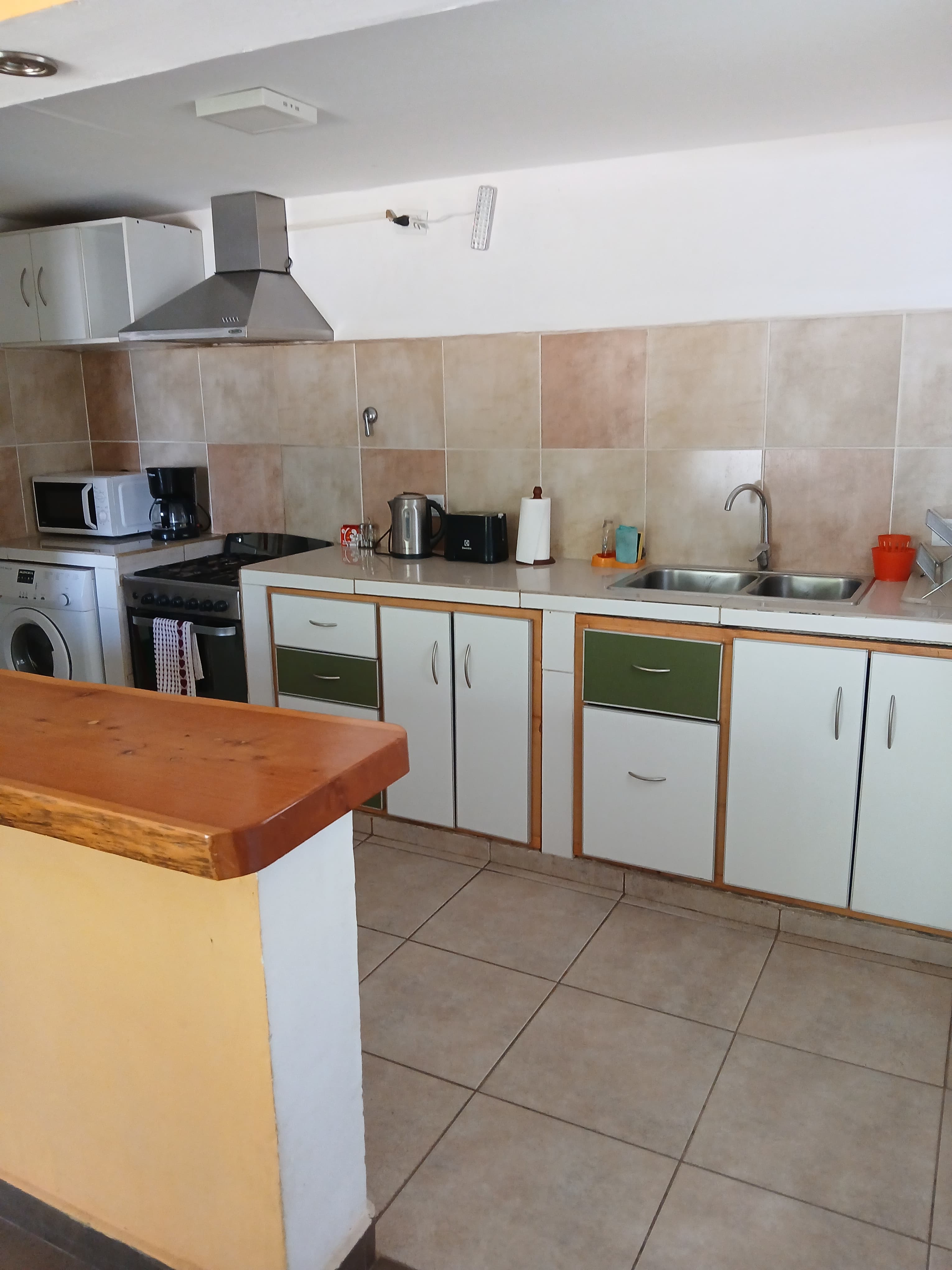 Cocina pequeña con gabinetes blancos y verdes, azulejos beige, fregadero y electrodomésticos compactos integrados.