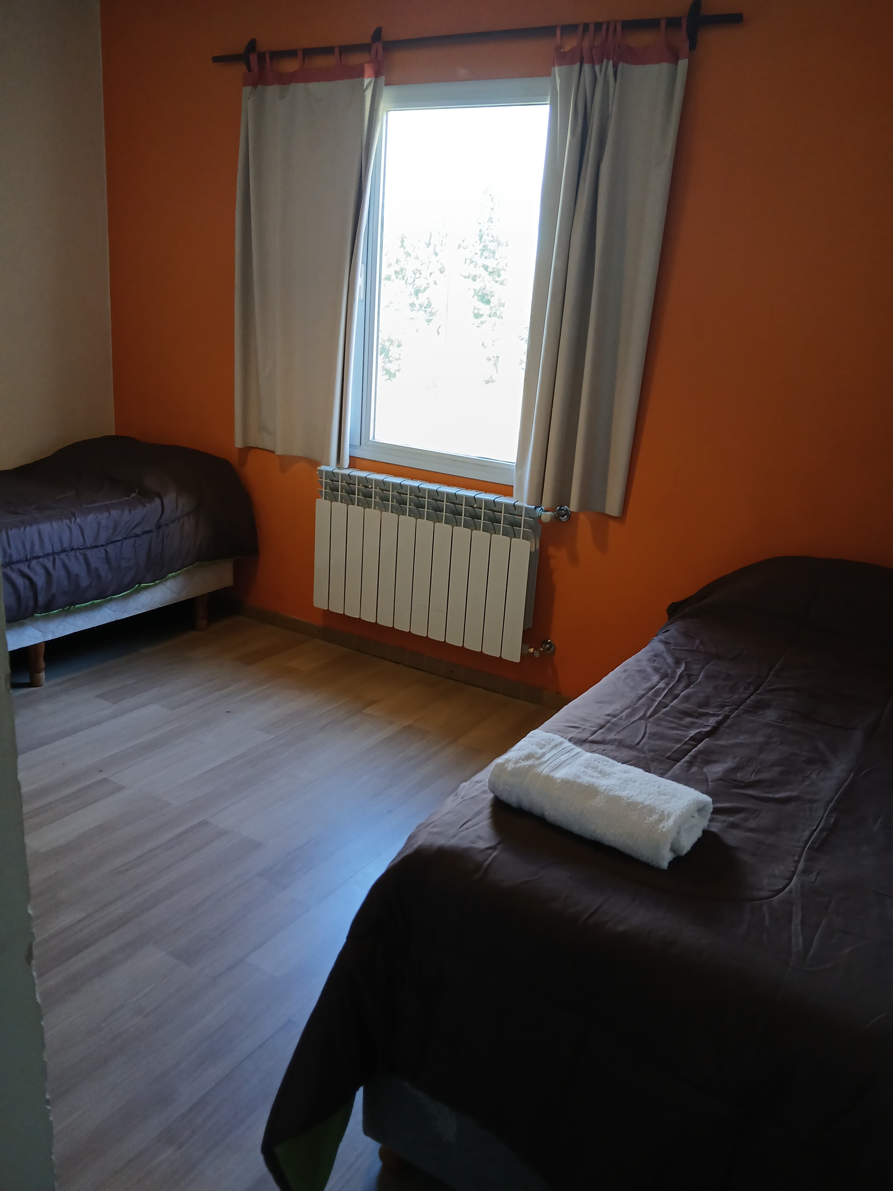 Dormitorio con paredes naranjas, dos camas con colchas marrones, ventana con cortinas y radiador blanco.
