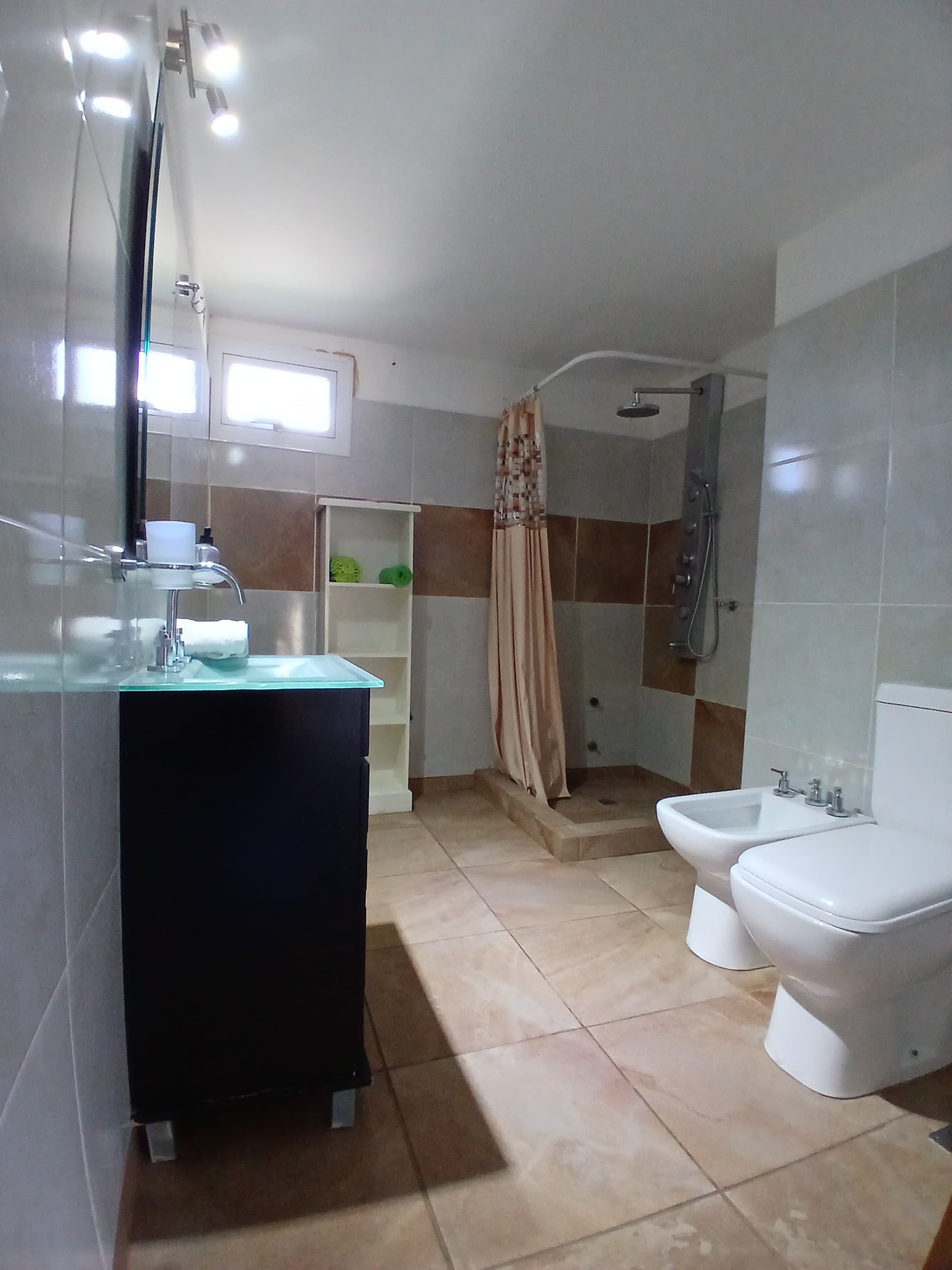 Baño moderno con inodoro blanco, bidé, ducha con cortina y un mueble de lavabo oscuro.