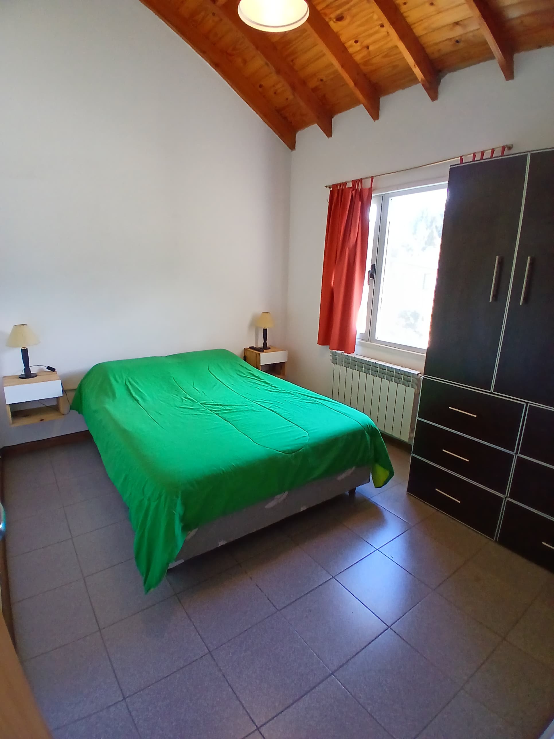 Habitación con cama verde, techo de madera inclinado, cortinas rojas y un armario oscuro.