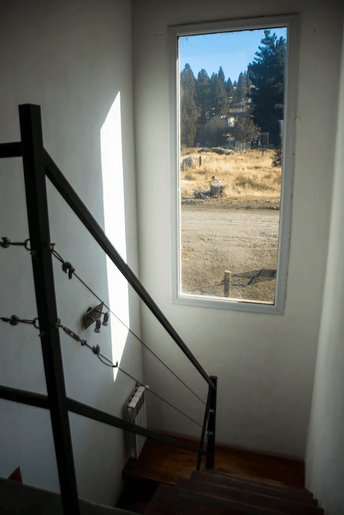 Escalera con barandilla negra y ventana con vistas a un camino rural y campo seco.