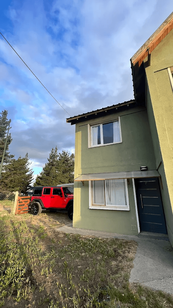 Casa verde de dos niveles con un Jeep rojo estacionado junto a unos pinos.
