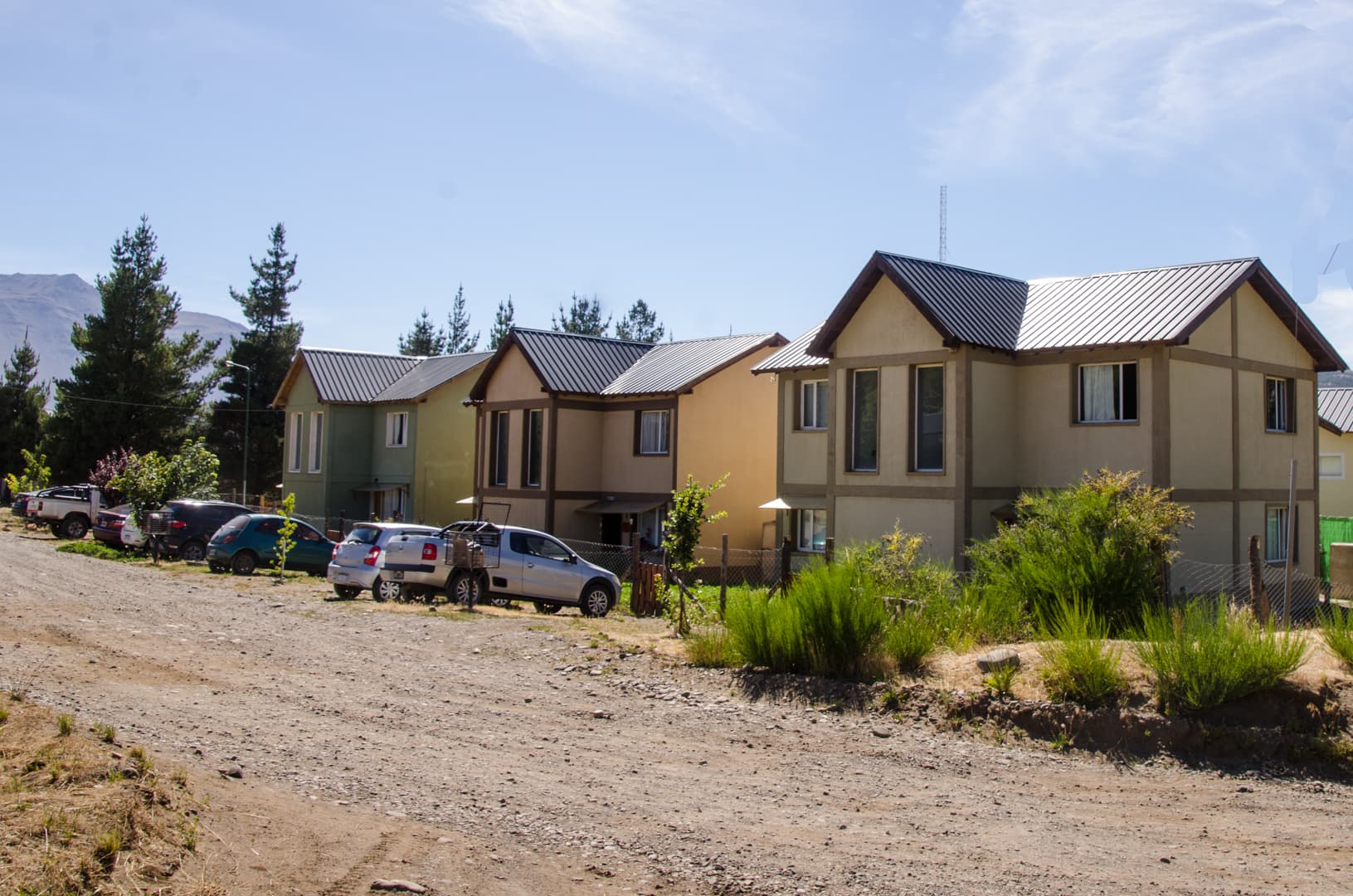 Casas modernas de dos pisos alineadas frente a un camino de tierra con autos estacionados.