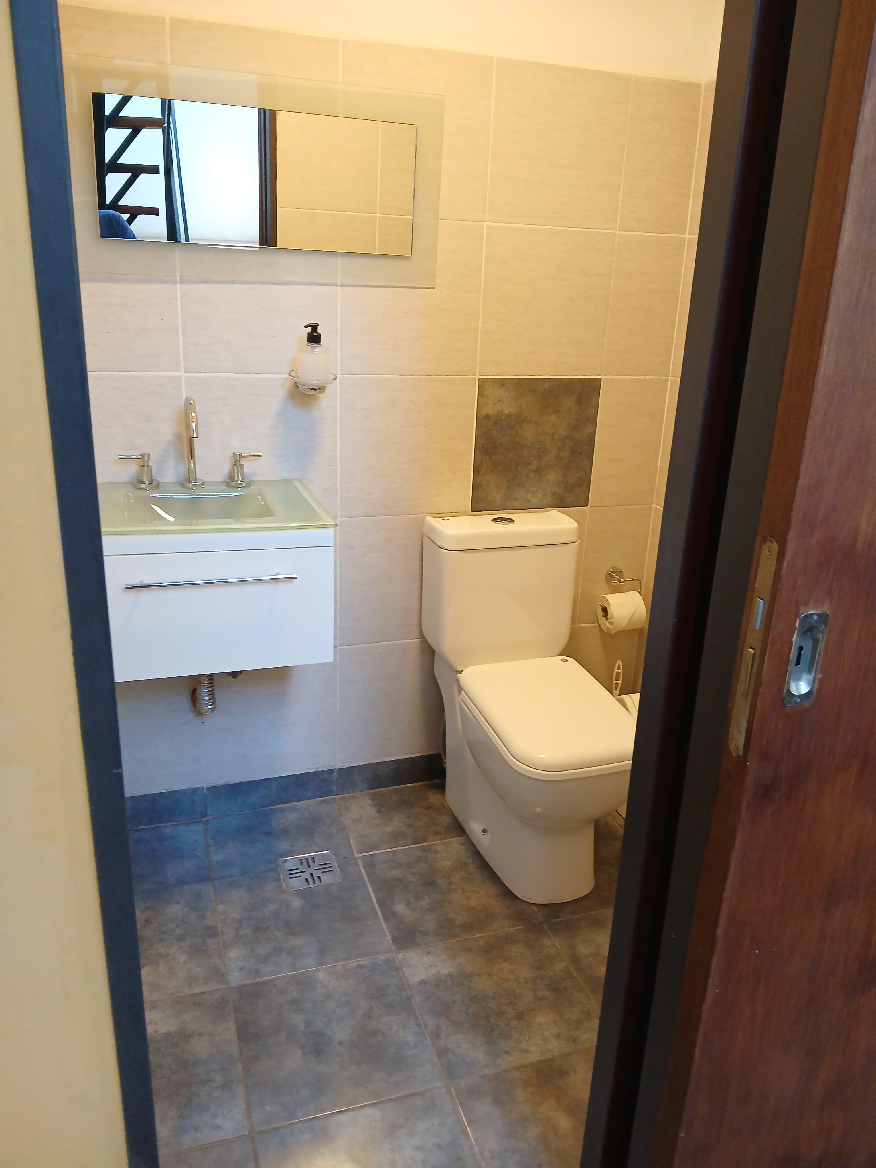 Baño moderno con inodoro blanco, mueble de lavabo, espejo y paredes de azulejos claros.
