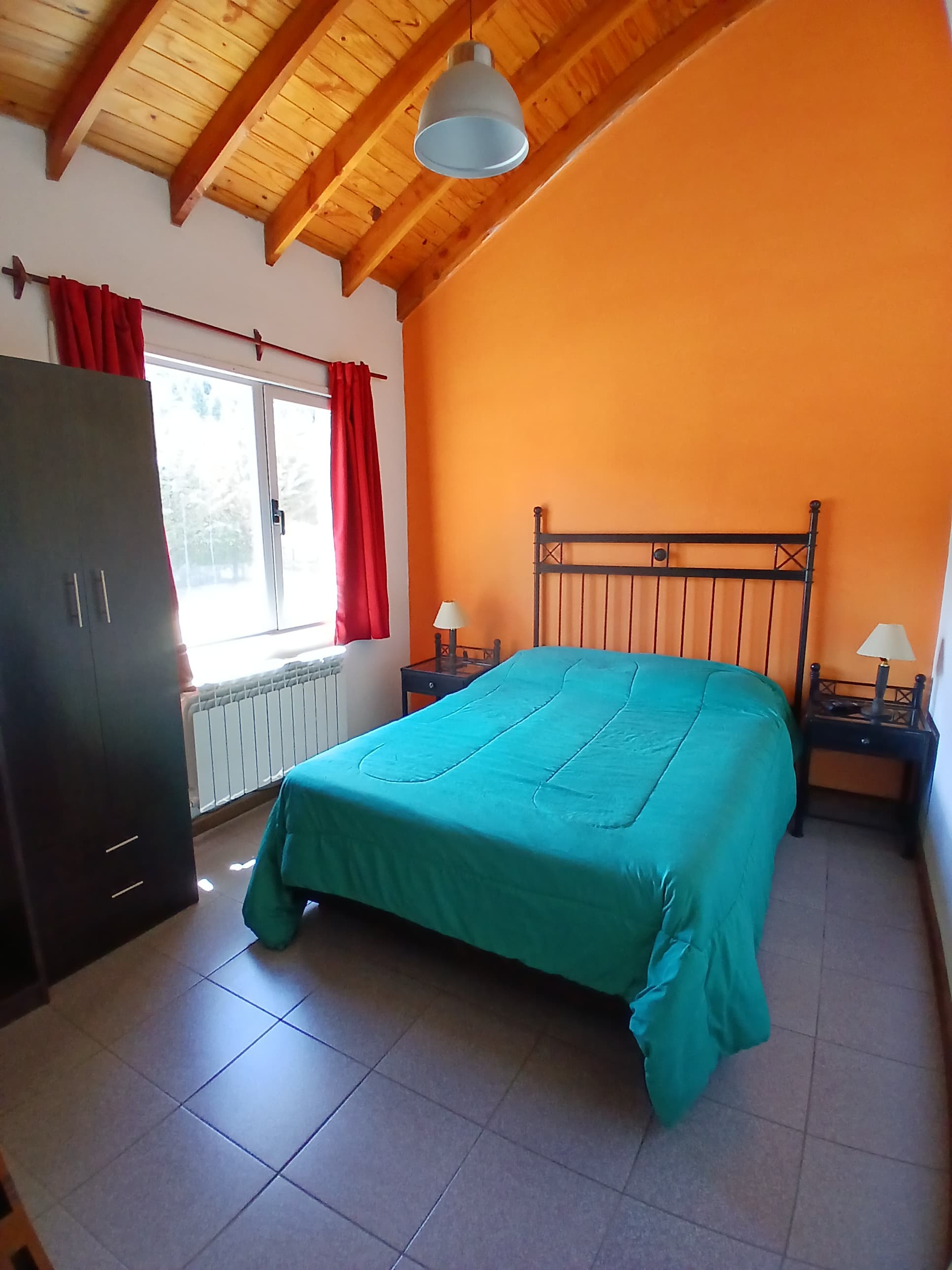 Dormitorio con pared naranja, cama verde, techo de madera rústico y ventana con cortinas rojas.