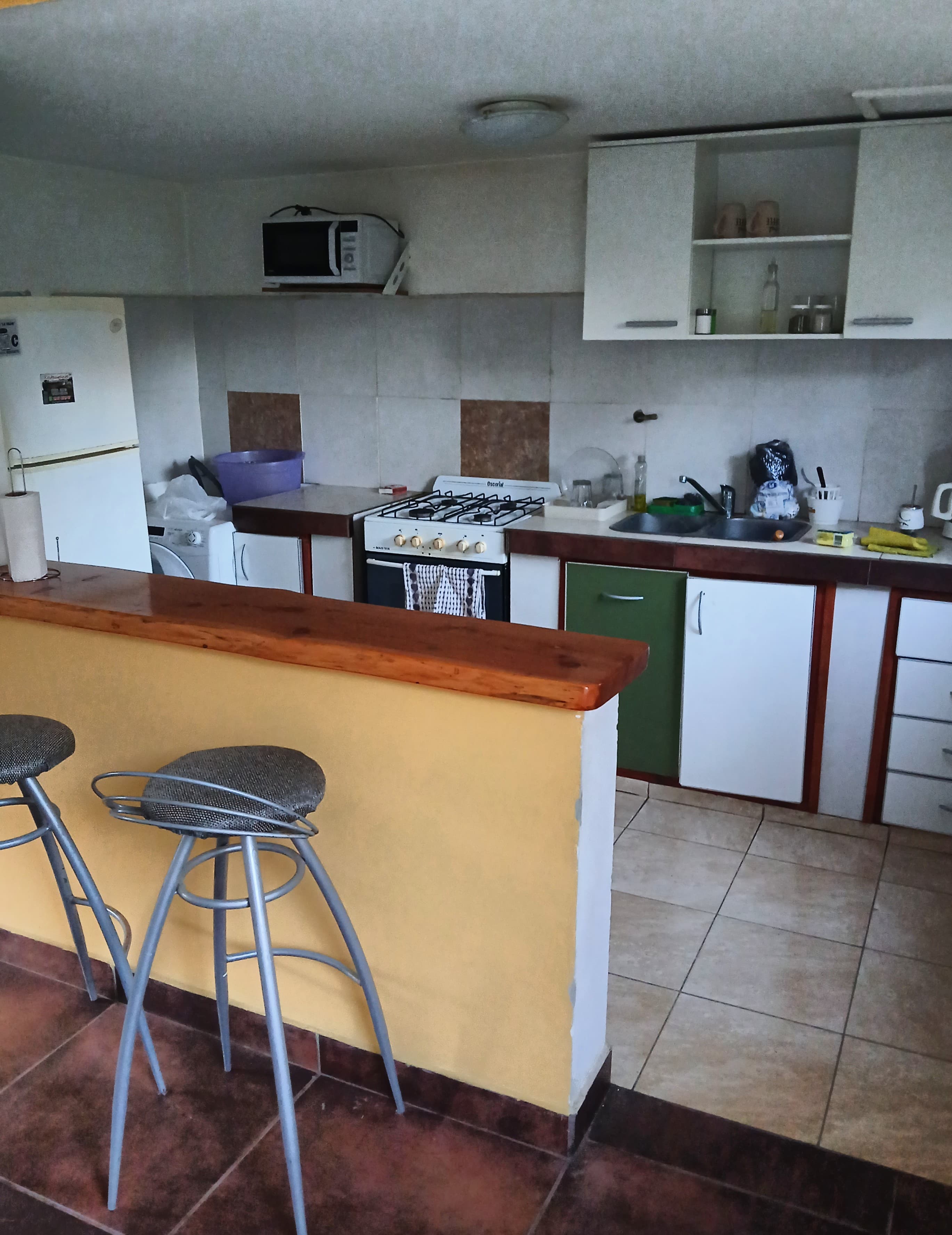 Cocina con barra amarilla, taburetes metálicos, estufa blanca, fregadero y gabinetes blancos y verdes.