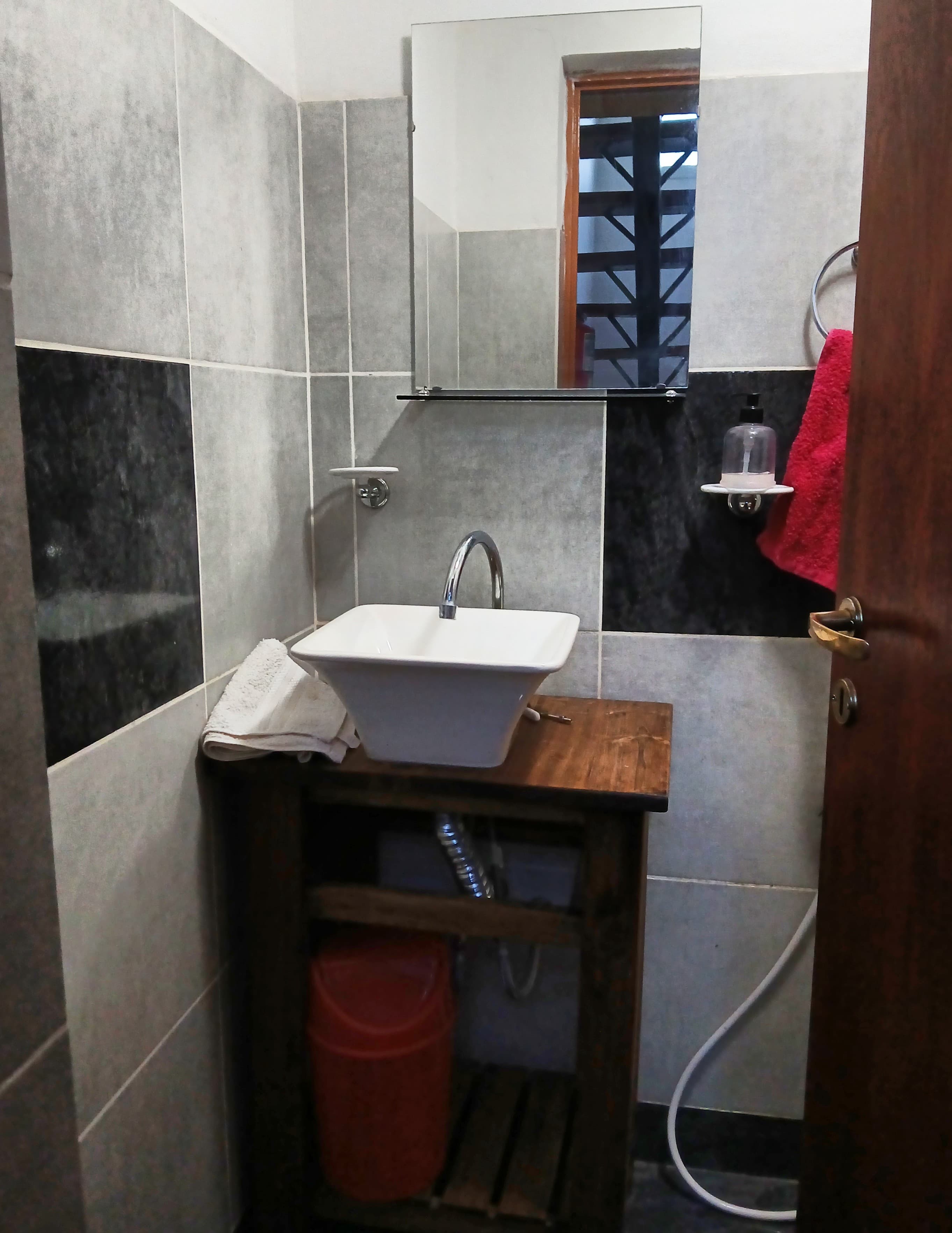 Lavabo blanco sobre mueble de madera, espejo y paredes con azulejos grises y negros.