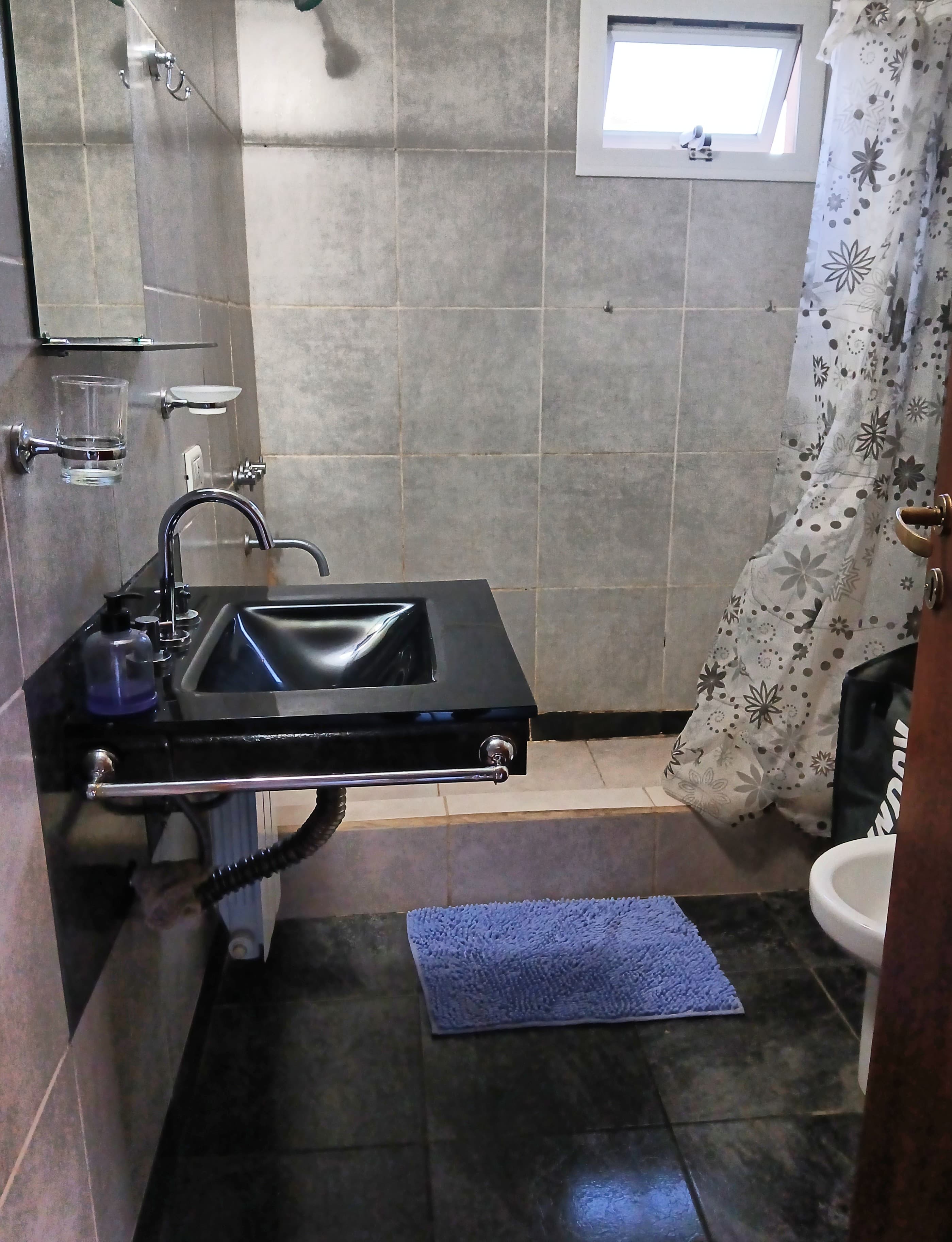 Baño con lavabo negro, paredes grises, alfombra azul y cortina de ducha con diseño floral.